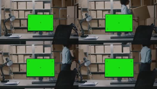 在仓库计算机与绿色模拟屏幕显示站在桌面上。在背景的货架上装满了纸箱和包裹的产品准备运输。高清在线视频素材下载