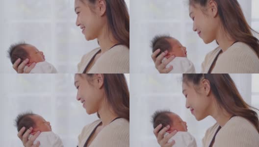 美丽迷人的亚洲妈妈抱着刚出生的宝宝，亲吻着宝宝的头，甜美可爱。快乐的妈妈和宝宝一起看着宝宝，带着爱微笑。母亲，婴儿，新生儿概念。慢动作高清在线视频素材下载