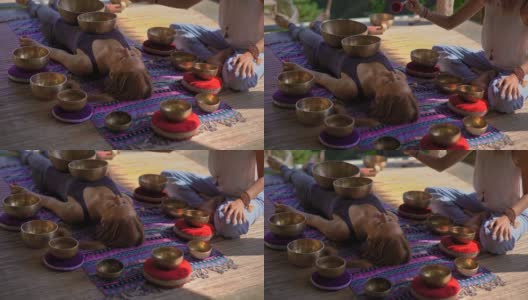 一名精通亚洲圣药的女子为一位年轻女性客户表演藏式碗治疗仪式的超慢镜头。用西藏唱碗冥想。他们在一个凉亭里冥想，背景是美丽的瀑布高清在线视频素材下载