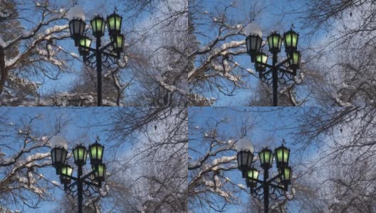 蓝蓝的天空下闪烁着绿光的街灯，黑色的树枝覆盖着雪花的剪影，街灯柱带有十字结构高清在线视频素材下载