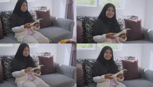 年长的亚洲穆斯林妇女戴着头巾，坐在沙发上休息，看书，看着相机，微笑着高清在线视频素材下载