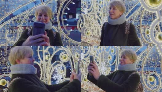 在圣诞节和新年期间，一个迷人的成熟女人，一名游客，在一个大城市用智能手机自拍和拍摄灯火通明的节日装饰。高清在线视频素材下载