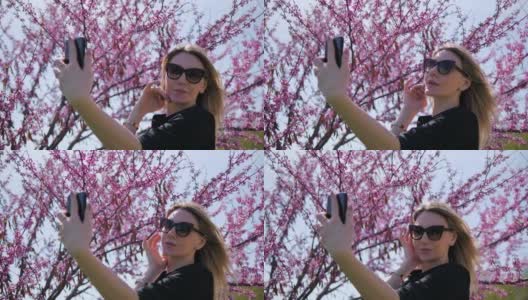 一位年轻美丽的女子在一棵盛开的粉红色樱花树的背景下自拍，欣赏自己高清在线视频素材下载