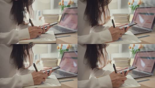 近距离的年轻亚洲女士自由职业聚焦手机聊天与同事工作财务写在笔记本电脑在客厅在家里。女学生在家在线学习，在家工作的概念。高清在线视频素材下载