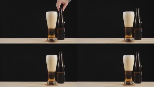 新鲜的啤酒。Hand把一个棕色的瓶子和一瓶美味的精酿啤酒放在一张黑色背景的木桌上。冰凉的新鲜啤酒，带有水滴，泡沫和泡沫。准备饮料。4 k高清在线视频素材下载