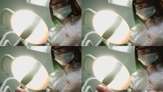 年轻女牙医戴着口罩，带着工具走近。牙科灯照进病人嘴里，站在病人身上，看着相机，牙医的脸高清在线视频素材下载