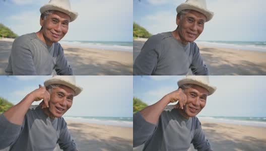 亚洲老人戴着帽子坐在沙滩上微笑着用手势打电话聊天。高清在线视频素材下载