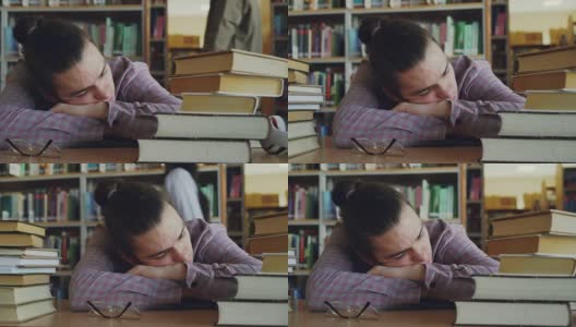 英俊的年轻男子坐在他的头在桌子上睡觉，在学校图书馆周围的大量书籍。他的眼镜放在他面前高清在线视频素材下载