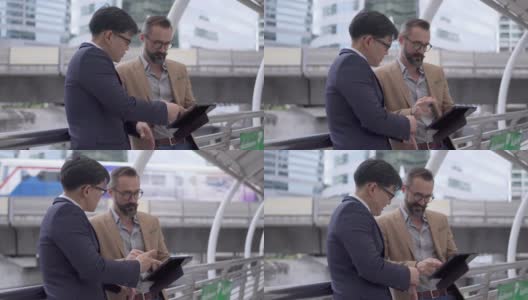 两个商务人士白种人与亚洲人同事伙伴关系讨论商业项目计划与使用数字平板电脑在城市。商务会议、技术、并购概念。高清在线视频素材下载