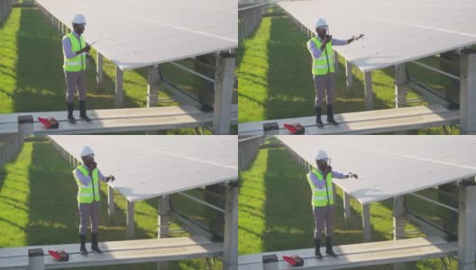 在太阳能发电厂安装太阳能电池板的过程中，非洲的年轻工程师穿着防护服和反光布爬上并站在结构上，通过无线电与团队联系高清在线视频素材下载