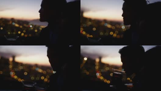 一个男人和女人站在屋顶上喝着鸡尾酒在夕阳下的城市灯光背后的特写高清在线视频素材下载