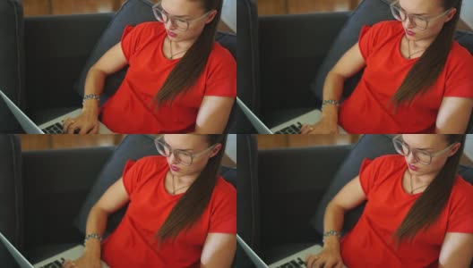 一位身穿红色连衣裙的黑发女子正坐在椅子上，在笔记本电脑的键盘上打字。远程雇佣，女性自由职业者，社交网络高清在线视频素材下载