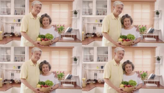 亚洲老夫妇感到幸福的微笑，拿着水果，看着镜头，而放松在厨房在家里。生活方式老年人家庭享受居家时光的概念。看着相机的人像。高清在线视频素材下载