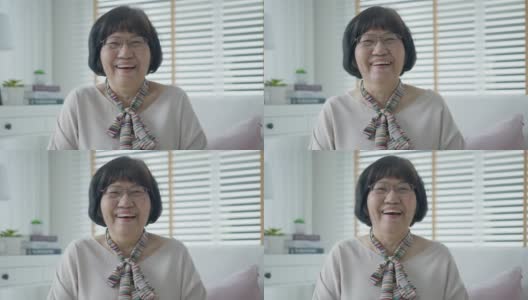 头像快乐健康的中年亚洲女人戴着眼镜坐在家里的沙发上。老夫人笑着，微笑着，看着客厅里的摄像机。屏幕视频通话截图。高清在线视频素材下载