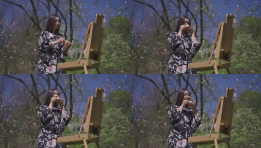 美丽苗条的亚洲女人在和服喝茶站在画架在阳光明媚的春天公园户外。这是一位才华横溢的女画家的肖像。美丽的概念。高清在线视频素材下载