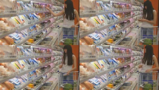 年轻女子推着购物车在超市的冷藏区购买乳制品或冷藏食品。一个女孩走到商店的冰箱前，从里面拿东西高清在线视频素材下载