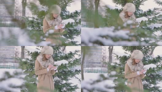 美丽的女人戴着皮草帽在冬天的公园里使用智能手机在下雪的天气下高清在线视频素材下载