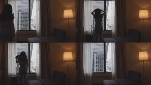 照片中年轻的女子带着她的行李在酒店的卧室里走着，站在窗边，看着美丽的城市风景，女子轻拂着她的头发享受假期旅行。慢动作高清在线视频素材下载