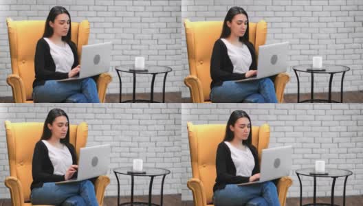 沉思的亚洲女人聊天用笔记本电脑坐在黄色的扶手椅上。由RED Raven 4k电影摄像机拍摄高清在线视频素材下载