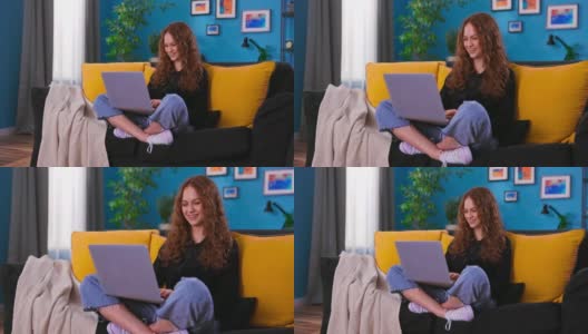 一个愉快的年轻女人坐在沙发上，把笔记本电脑放在膝盖上，浏览互联网，社交网络，网购。十几岁的女孩坐在沙发上使用笔记本电脑高清在线视频素材下载