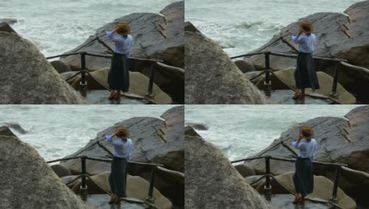 一个戴着棕色帽子的女孩站在多石的河岸上看着海浪高清在线视频素材下载