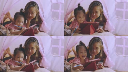 一张专注于亚洲妈妈和可爱的女儿躺在家里粉色帐篷的地板上，在使用数字技术的平板电脑上写着快乐和快乐的时刻。高清在线视频素材下载