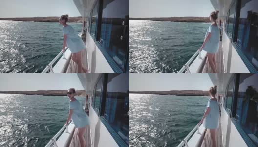 一个年轻美丽的白人女人在一个游艇上的肖像在蓝色的裙子和太阳镜看着一个荒岛。假期的概念，自由，阳光夏日。背景是蓝色的海洋。steadycam高清在线视频素材下载