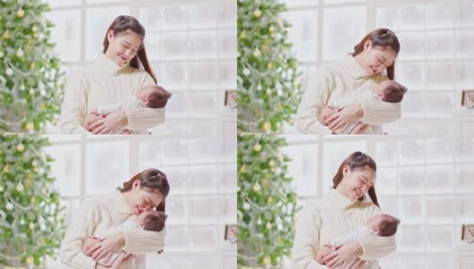 美丽的亚洲妇女抱着新生的婴儿，亲吻坐在白色舒适的家里的床上，背景是圣诞树。快乐的宝宝睡在妈妈的怀里，又安全又舒适高清在线视频素材下载