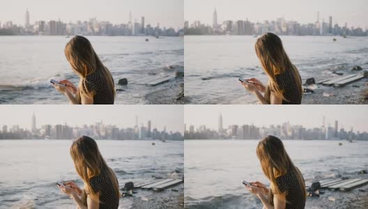 在河滩上使用智能手机应用程序，在4K日落下看到纽约市的天际线高清在线视频素材下载