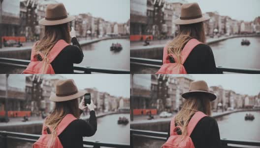 戴着帽子的年轻女孩在风景优美的河边拍照。一个留着长发背着红色背包的女孩在她的博客上分享照片。4 k高清在线视频素材下载