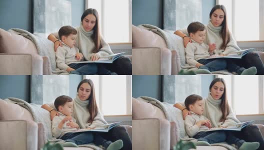 年轻漂亮的妈妈坐在你家客厅的沙发上给儿子读故事，学着读小男孩高清在线视频素材下载