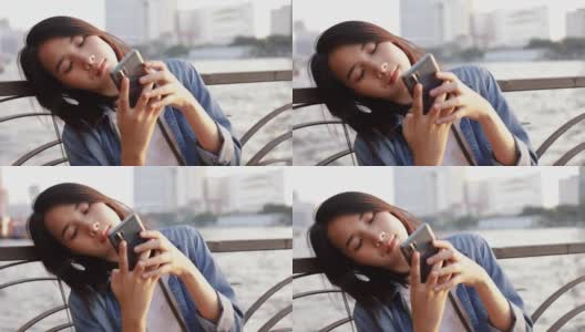 快乐的亚洲女性使用智能手机与她的朋友聊天，而坐在湄南河在泰国在美丽的日落。高清在线视频素材下载