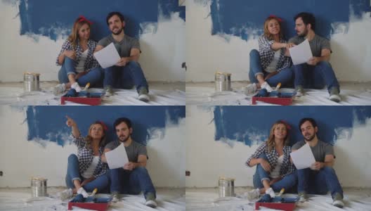 一对幸福的夫妇坐在地板上看着纸上的设计在家庭装修期间高清在线视频素材下载