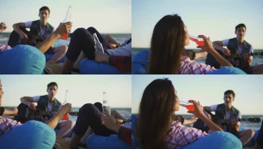 一群朋友坐在沙滩上的安乐椅上，喝着鸡尾酒和啤酒，欢呼雀跃，听着朋友在一个夏日傍晚的夕阳下弹吉他。Slowmotion拍摄高清在线视频素材下载