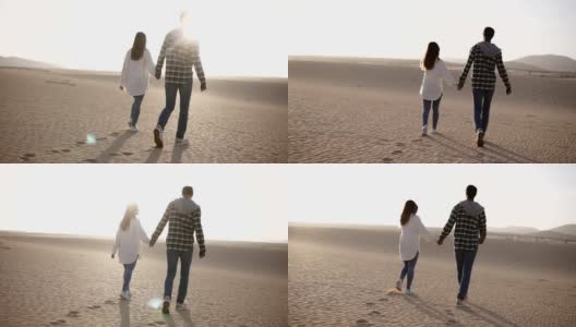 这是罕见的一对情侣在阳光下和沙漠里的蓝天下牵手浪漫地散步。两个年轻的恋人，穿着休闲的衣服，手牵着手走在沙漠的沙子上。镜头耀斑高清在线视频素材下载