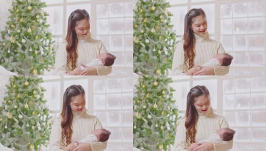 美丽的亚洲妇女抱着新生儿坐在白色舒适的家里的床上，背景是圣诞树。快乐的宝宝睡在妈妈的怀里，又安全又舒适。母婴概念高清在线视频素材下载