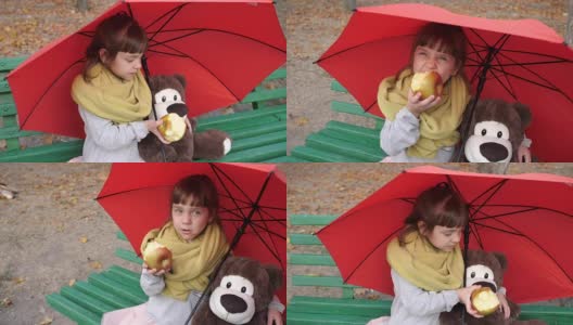 小女孩和玩具朋友坐在公园的一把伞下的长椅上吃红苹果高清在线视频素材下载