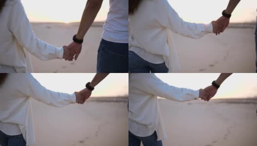 镜头中，这对牵手的情侣在阳光下和沙漠中的蓝天下浪漫地散步。两个年轻的恋人穿着休闲的衣服漫步在沙漠的沙滩上。景观上的背景高清在线视频素材下载