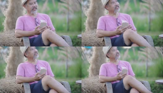 笑容满面的亚洲灰胡子成熟男子坐在户外的花圃里，用手机上的在线约会app聊天。快乐的退休男人穿着粉色衬衫，戴着眼镜，日落时戴着帽子。普通情人节新概念高清在线视频素材下载