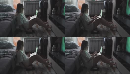 在一个多云的清晨，一名年轻女子正坐在电视机前的地毯上用笔记本电脑工作。她仰靠在摊开的沙发上。在家工作流行病商务距离高清在线视频素材下载