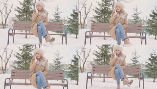 美丽的女人戴着皮草帽坐在长椅上，在冬天的城市公园里使用智能手机在下雪的天气里，慢动作高清在线视频素材下载
