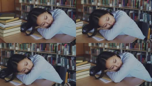 年轻美丽疲惫的亚洲女学生坐在书桌上，她的头在它上，她是睡在教科书周围的堆在大大学图书馆高清在线视频素材下载