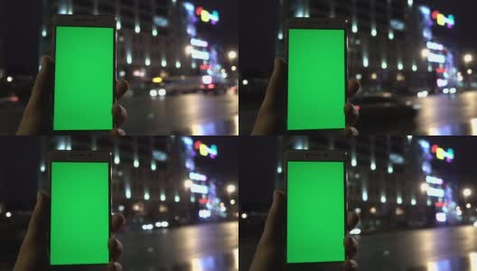 男子手握白色智能手机，在夜城灯光散景的背景下。绿色的屏幕。高清在线视频素材下载