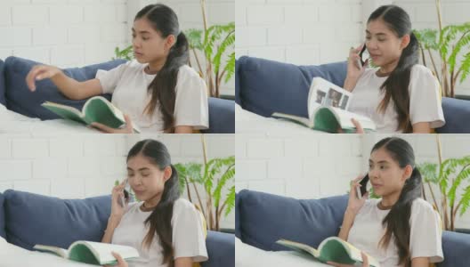 年轻美丽的亚洲女人谈论智能手机与幸福坐在沙发上看书，人在电话，生活方式高清在线视频素材下载