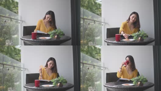 一位亚洲华裔美女中午在她的阳台上看书，放松，享受她的数字排毒时间，喝着咖啡，吃着饼干当点心高清在线视频素材下载