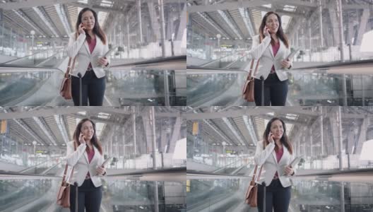 机场商务交流亚洲女商人，在机场出差，带着行李和肩袋在自动扶梯上与顾客交谈，低角度手持前视高清在线视频素材下载