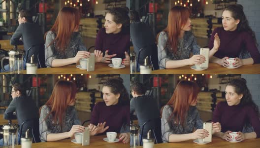 两个漂亮的女人穿着休闲衣服，在咖啡馆里聊着天，拿着咖啡杯和智能手机。很好的咖啡馆内部，顾客和家具都可以看到。高清在线视频素材下载