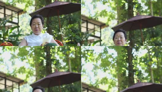 亚洲资深妇女喝咖啡快乐在绿色葱郁的自然咖啡店高清在线视频素材下载