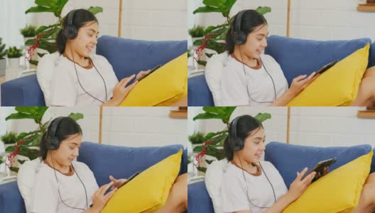 年轻美丽的亚洲女人坐在家里客厅的沙发上，用数码平板电脑和耳机听音乐，人和科技，Z一代的生活方式高清在线视频素材下载