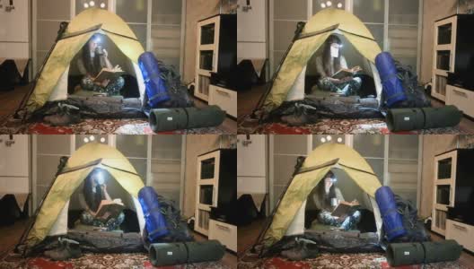 一个女孩戴着医用口罩在一间公寓的露营帐篷里看书。闭关锁国则在家里摧毁。冠状病毒大流行。怀念旅行和远足高清在线视频素材下载